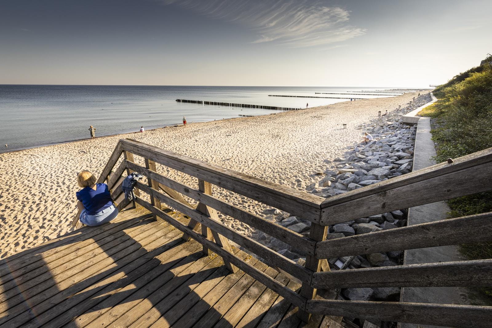 Holztreppe führt zu weitem Sandstrand und ruhiger Küste bei klarem Himmel.
