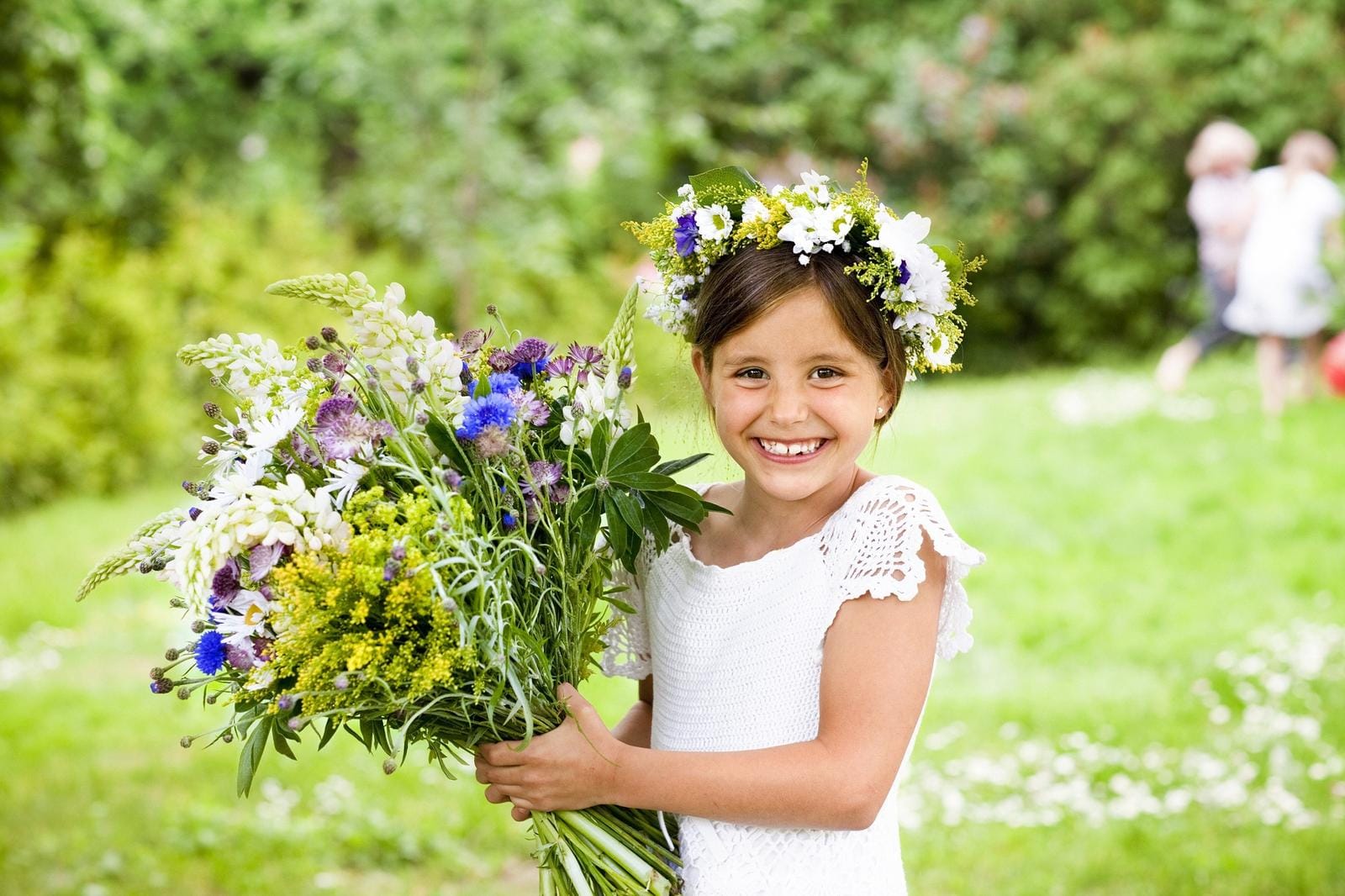 Ein Mädchen in weißem Kleid mit Blumenkranz lächelt, hält bunte Blumen im Garten.