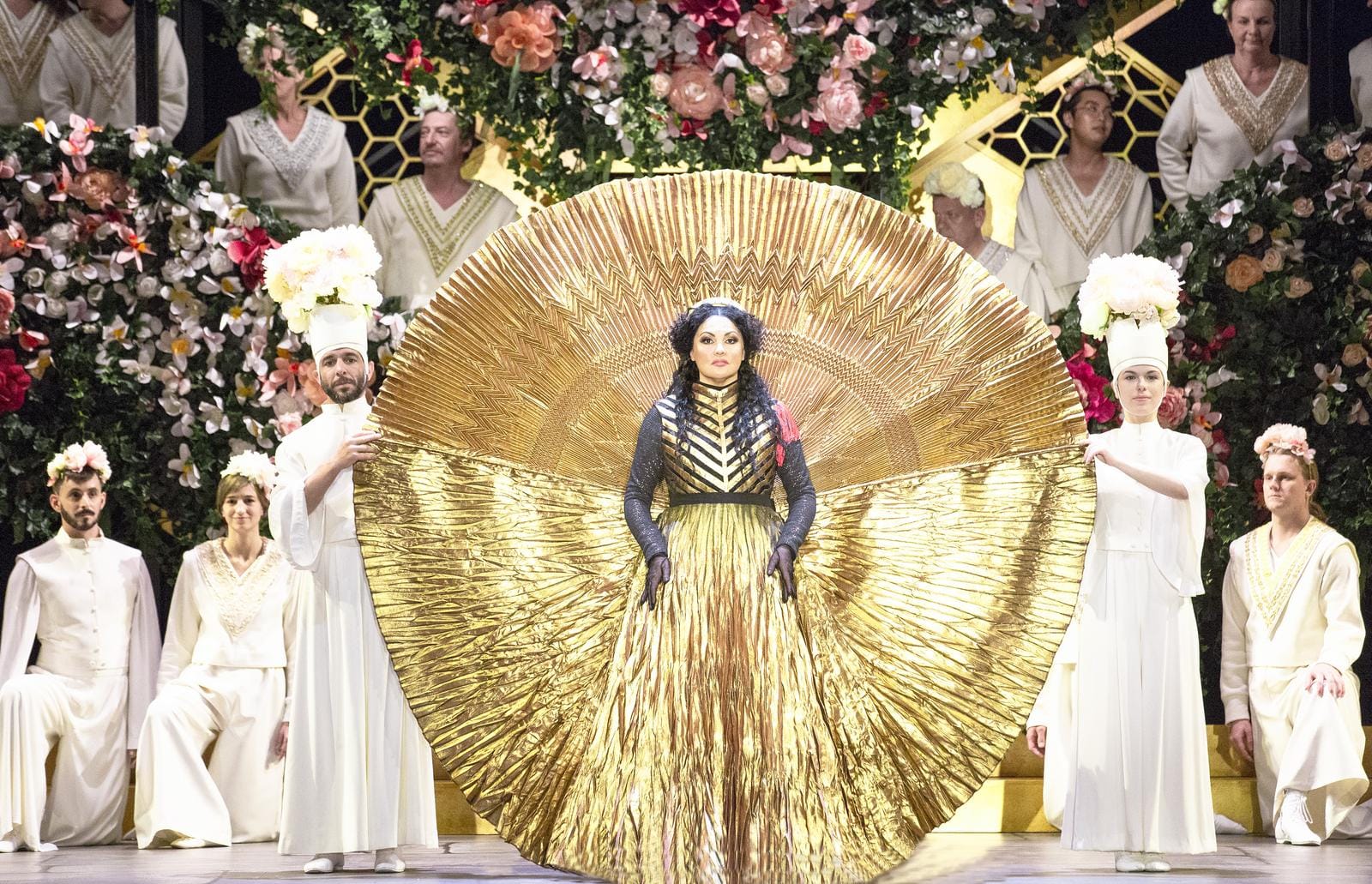 Opulentes Bühnenbild mit goldener Robe und Blütenkränzen vor floraler Kulisse.