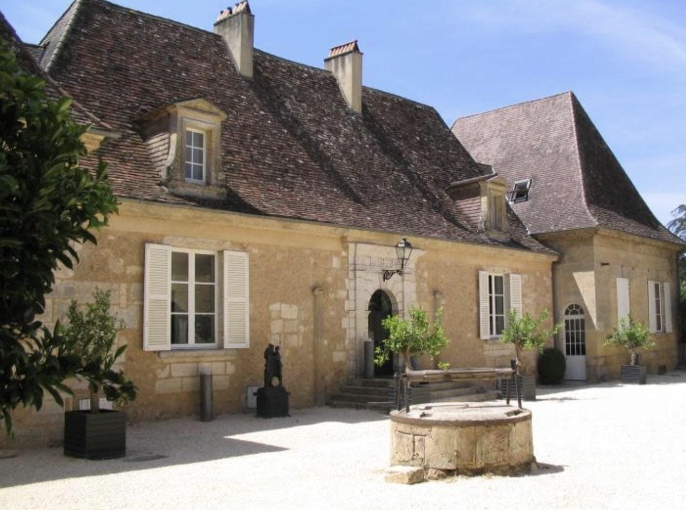 Historisches Steinhaus mit Schieferdach, weißen Fensterläden und Innenhof mit Brunnen.