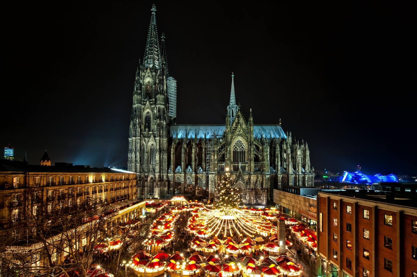 Kölner Dom bei Nacht mit beleuchtetem Weihnachtsmarkt, leuchtende Stände und Tannenbaum.