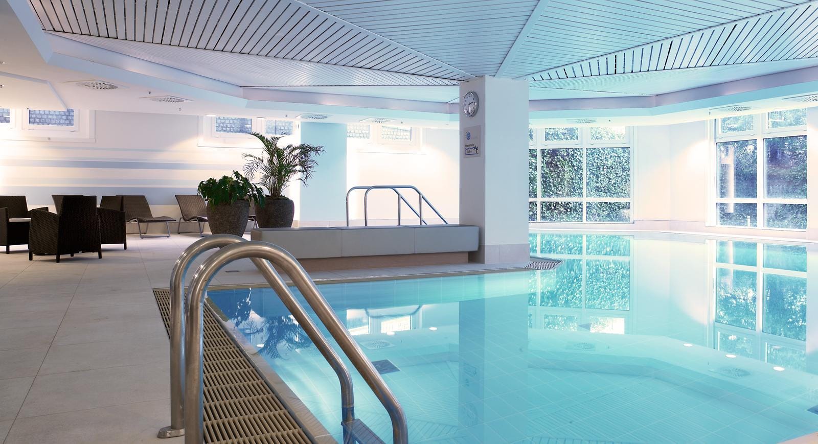 Helles Hallenbad mit modernen Liegestühlen, Pflanzen und großen Fenstern.