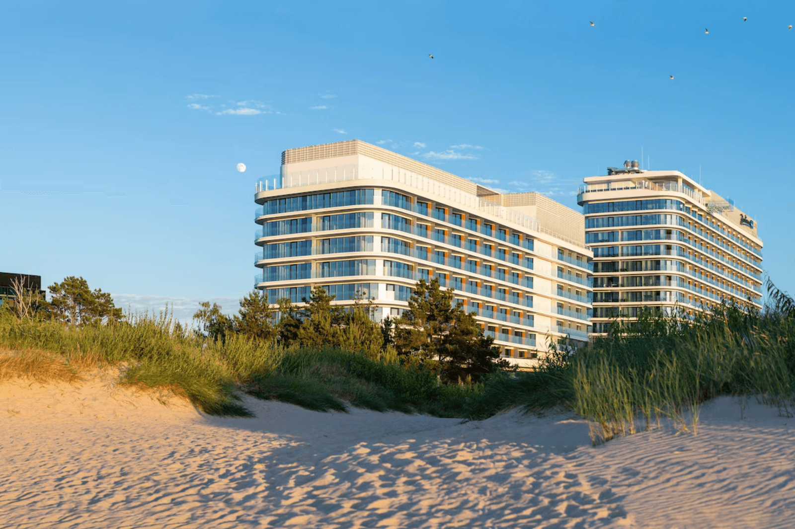 Modernes Strandhotel mit Glasfassade, hinter Dünen und blauem Himmel gelegen.