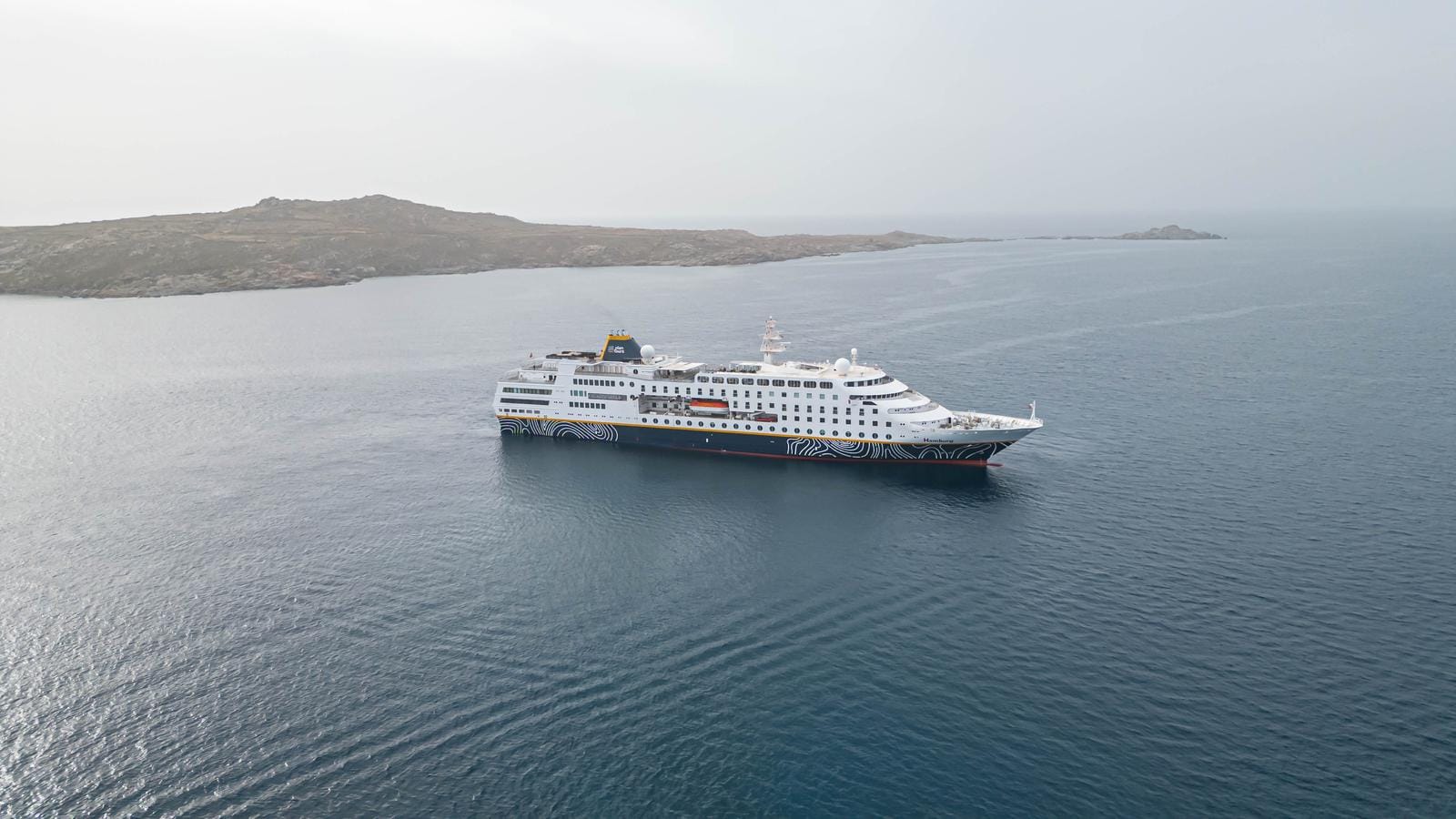 Kreuzfahrtschiff in ruhiger See vor felsiger Inselkulisse unter bewölktem Himmel.