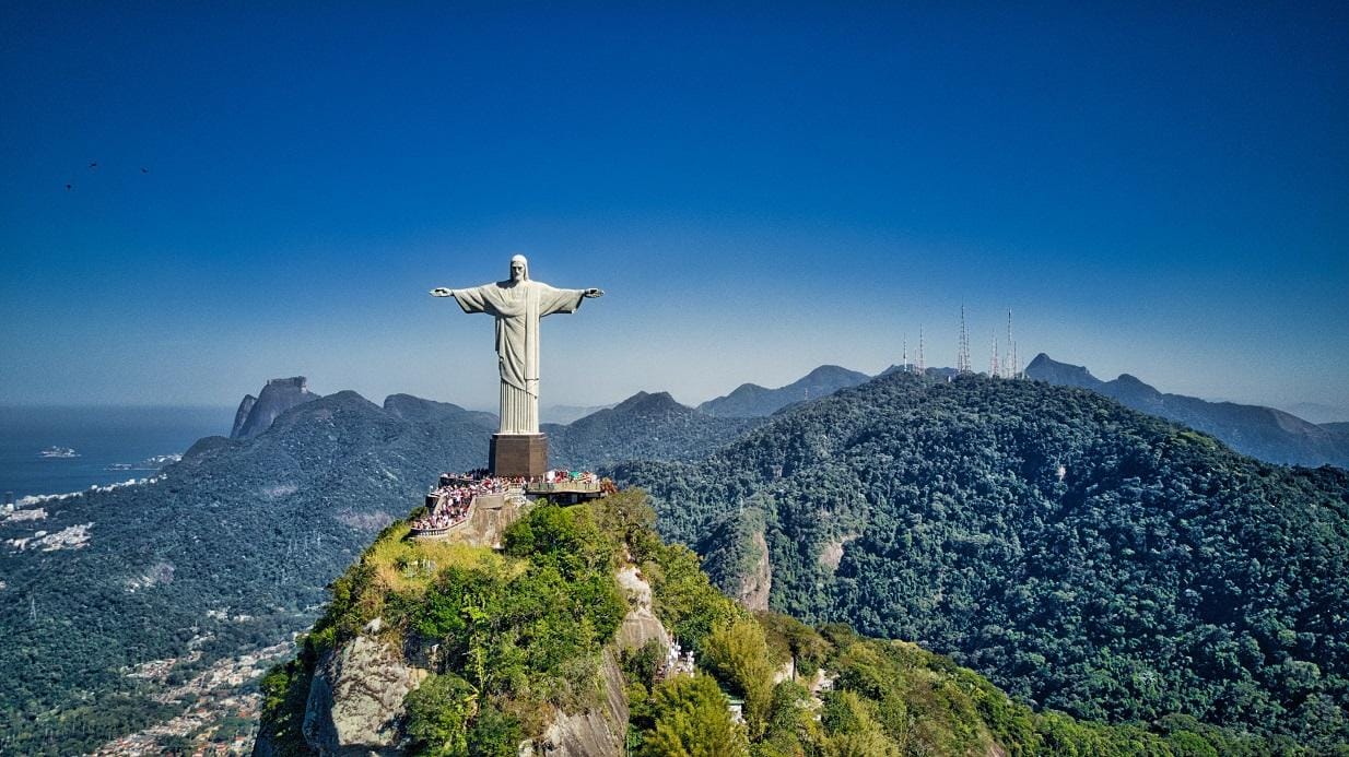 Christusstatue in Rio de Janeiro auf Berg, umgeben von grünen Wäldern und blauem Himmel.