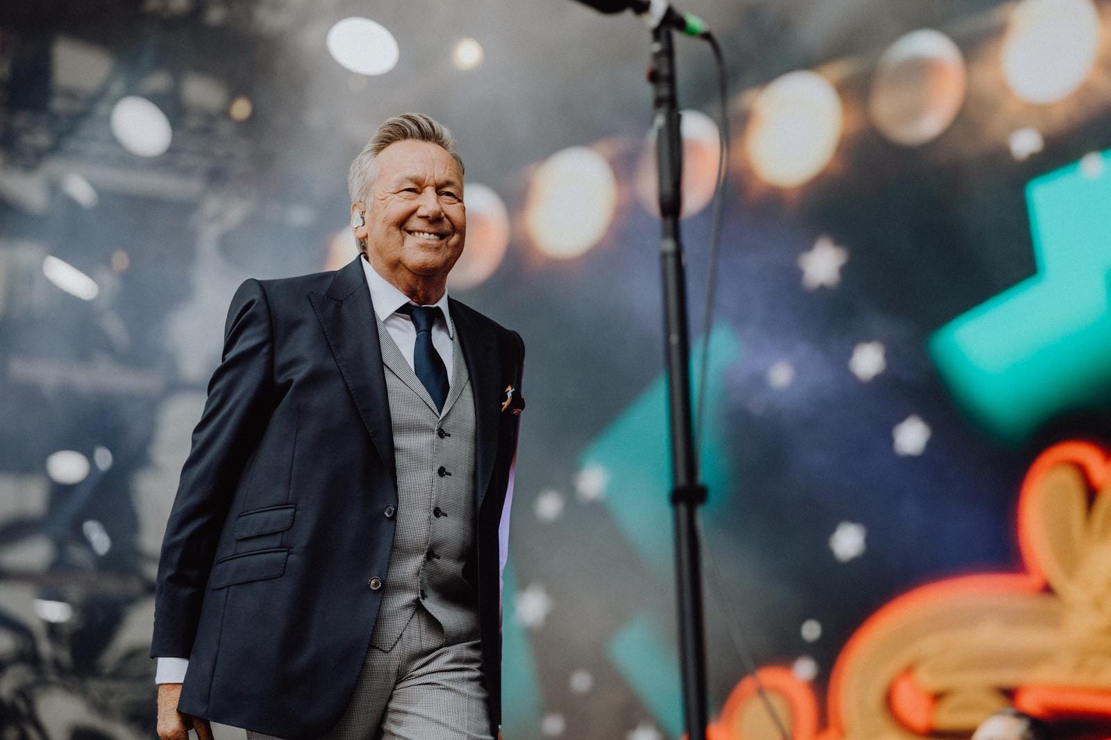 Ein lächelnder Mann im Anzug auf einer Bühne mit buntem, unscharfem Hintergrund.