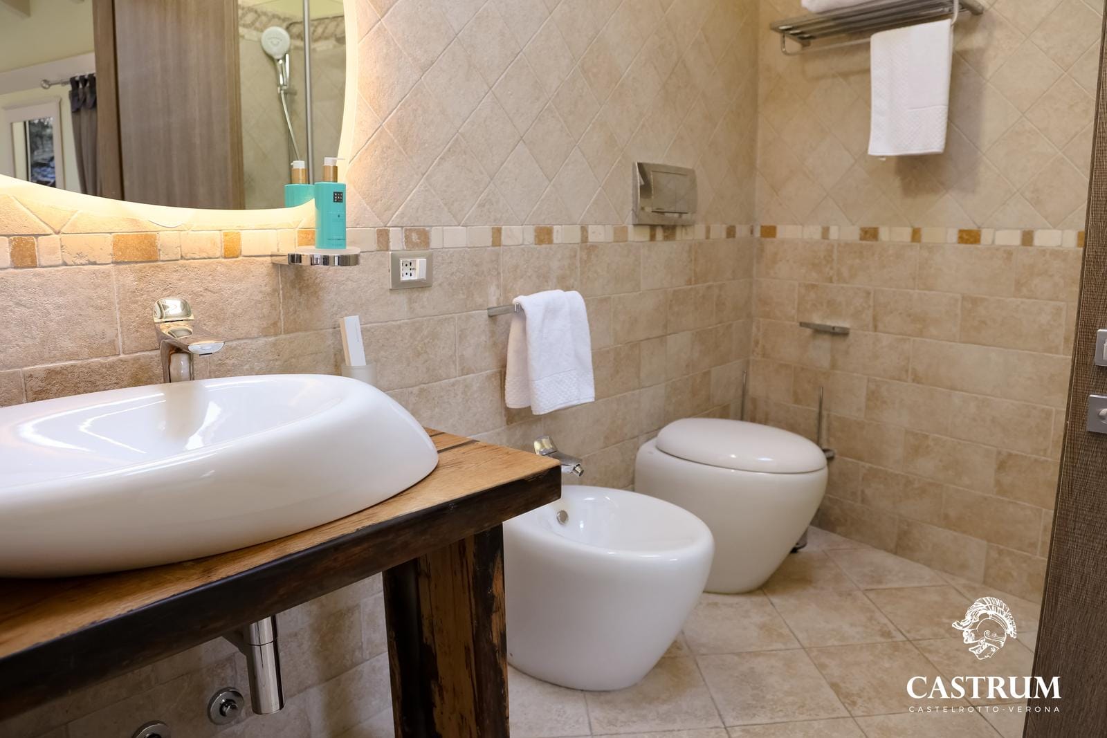 Modernes Hotelbad mit beigen Fliesen, weißem Waschbecken, Bidet und eleganter Beleuchtung.