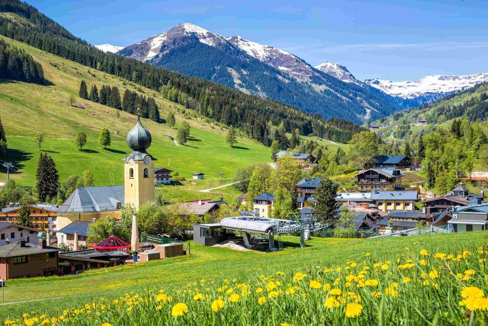 Dorf in den Alpen, Österreich, grüne Wiesen, blühende Löwenzahn, schneebedeckte Berge.