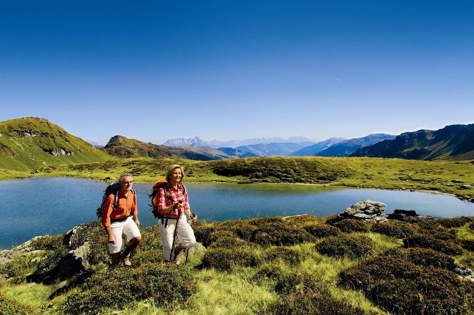 Zwei Wanderer vor einem klaren Bergsee in alpiner Landschaft bei strahlend blauem Himmel.