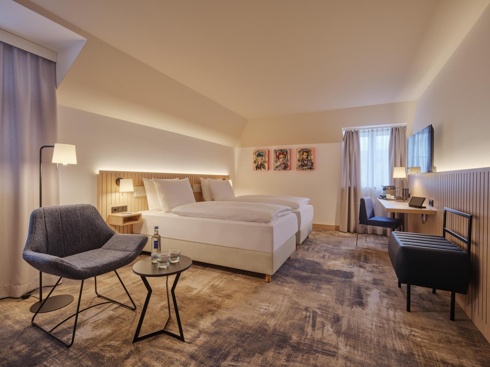Modernes Hotelzimmer mit Doppelbett, Sesseln, Schreibtisch, schlichter Dekor im warmen Licht.