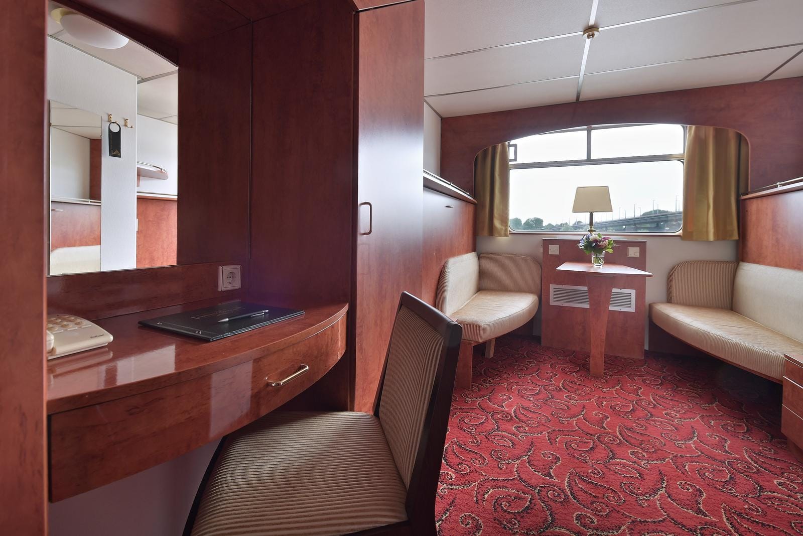 Gemütliches Hotelzimmer mit Holzmöbeln, rotem Teppich und Blick aus großem Fenster.