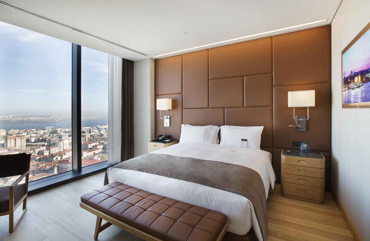 Modernes Hotelzimmer mit großem Bett, Lederrückenwand, Stadtblick, hellem Holzfußboden.