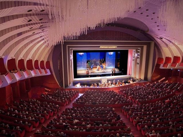 Opernhaus mit geschwungener Decke, roter Bestuhlung und beleuchteter Bühne.