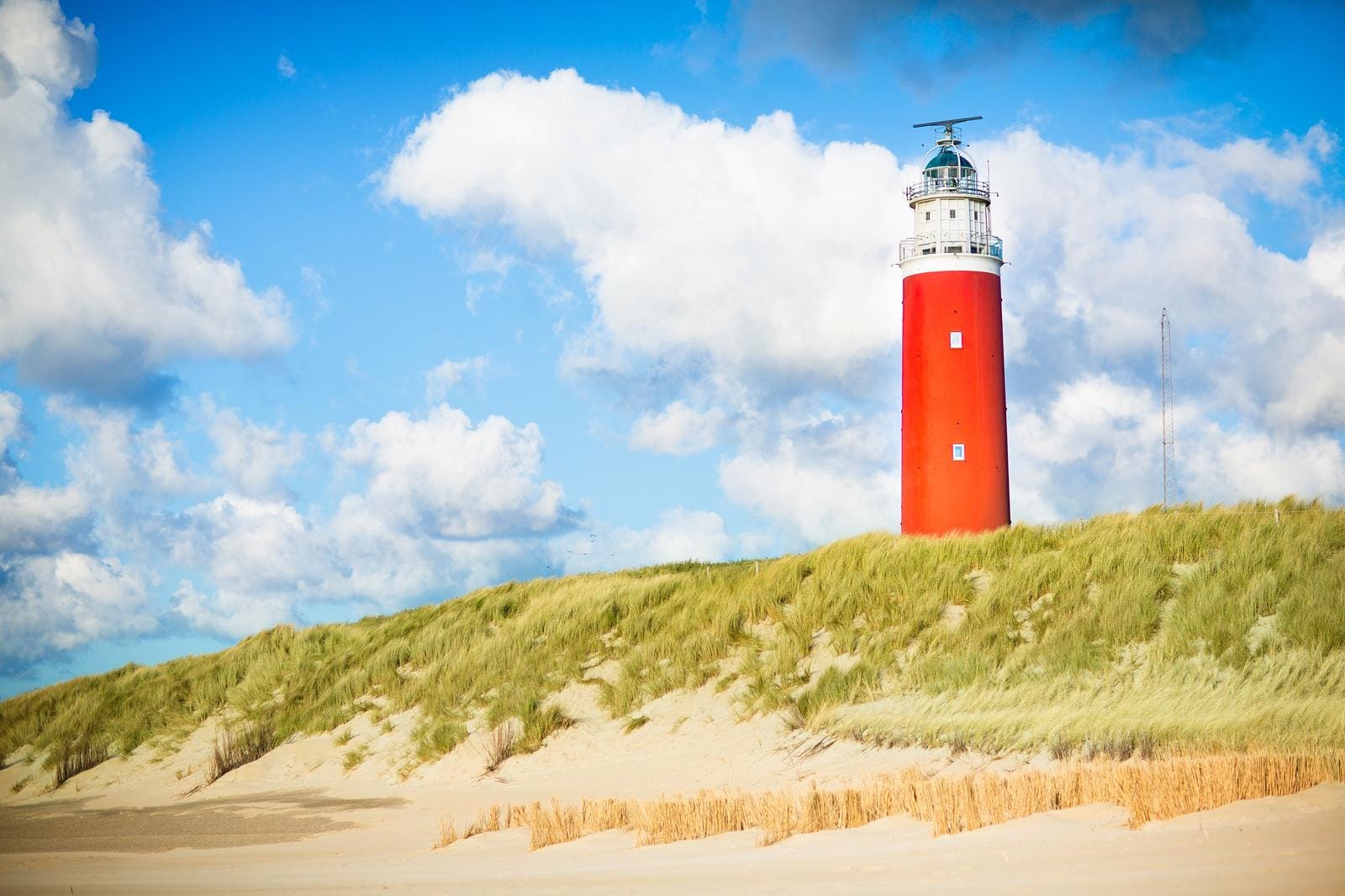 Roter Leuchtturm auf Dünen im Sonnenschein unter blauem Himmel mit weißen Wolken.
