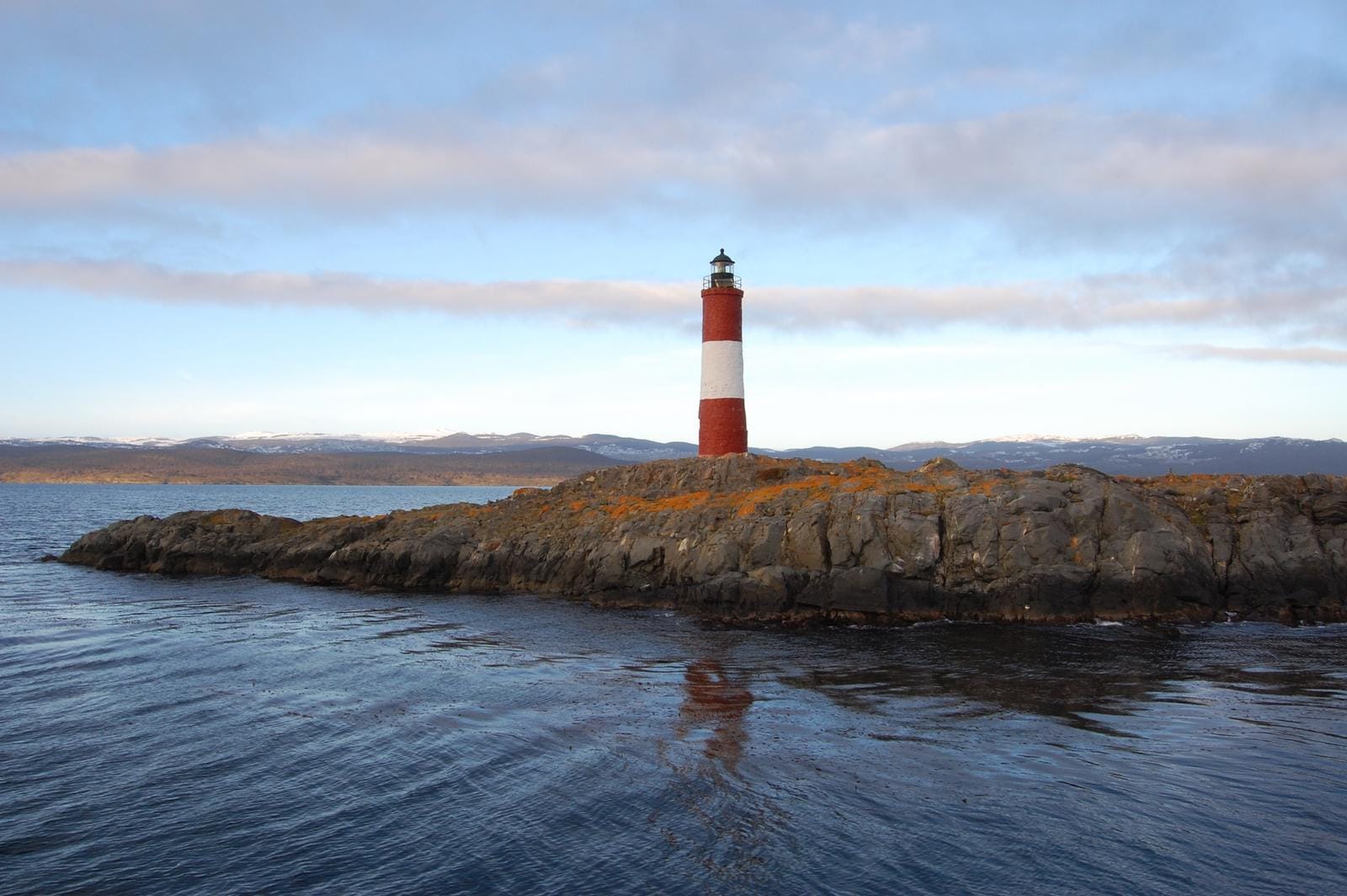 Rot-weißer Leuchtturm auf Felseninsel im Meer, im Hintergrund Berge und blauer Himmel.