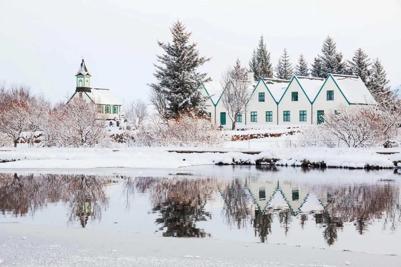 Verschneite Kirche und Gebäude in Thingvellir, Island, spiegeln sich im Wasser.
