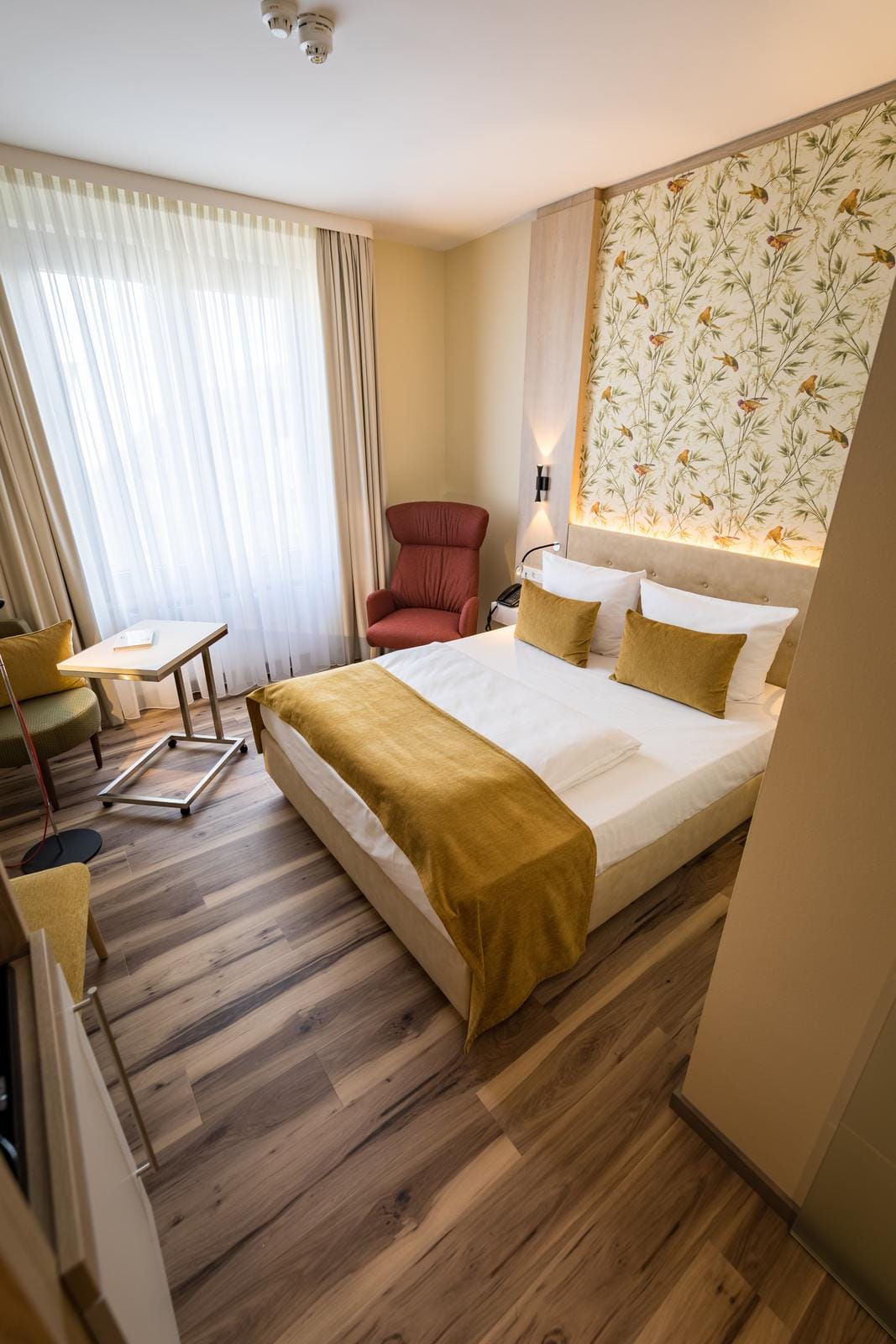 Helles Hotelzimmer mit floralem Tapetendesign, Holzboden und gemütlichen roten Sesseln.