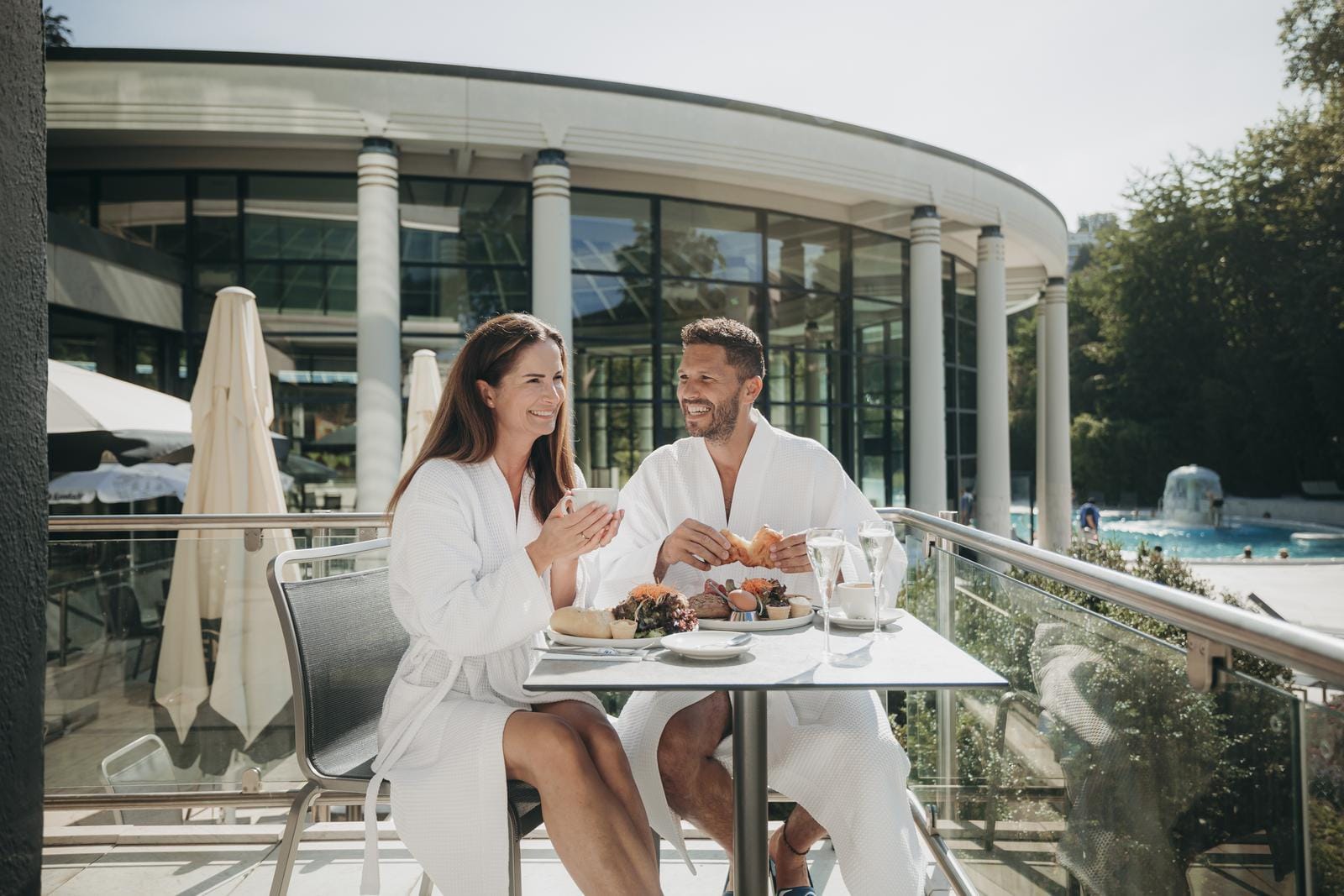 Paar frühstückt entspannt im Bademantel auf sonniger Hotelterrasse mit Poolblick.