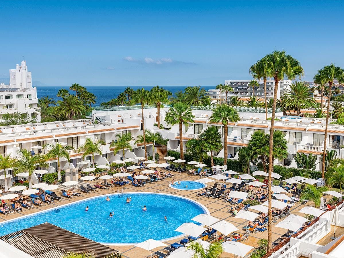Hotelpool mit Liegestühlen, Palmen, mediterranen Gebäuden und Meerblick bei sonnigem Himmel.