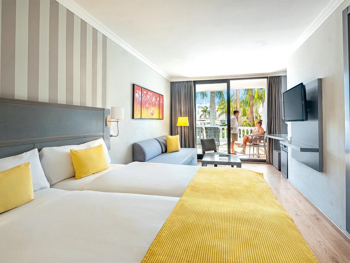 Modernes Hotelzimmer mit zwei Betten, gelben Akzenten und Balkon mit Blick auf Palmen.