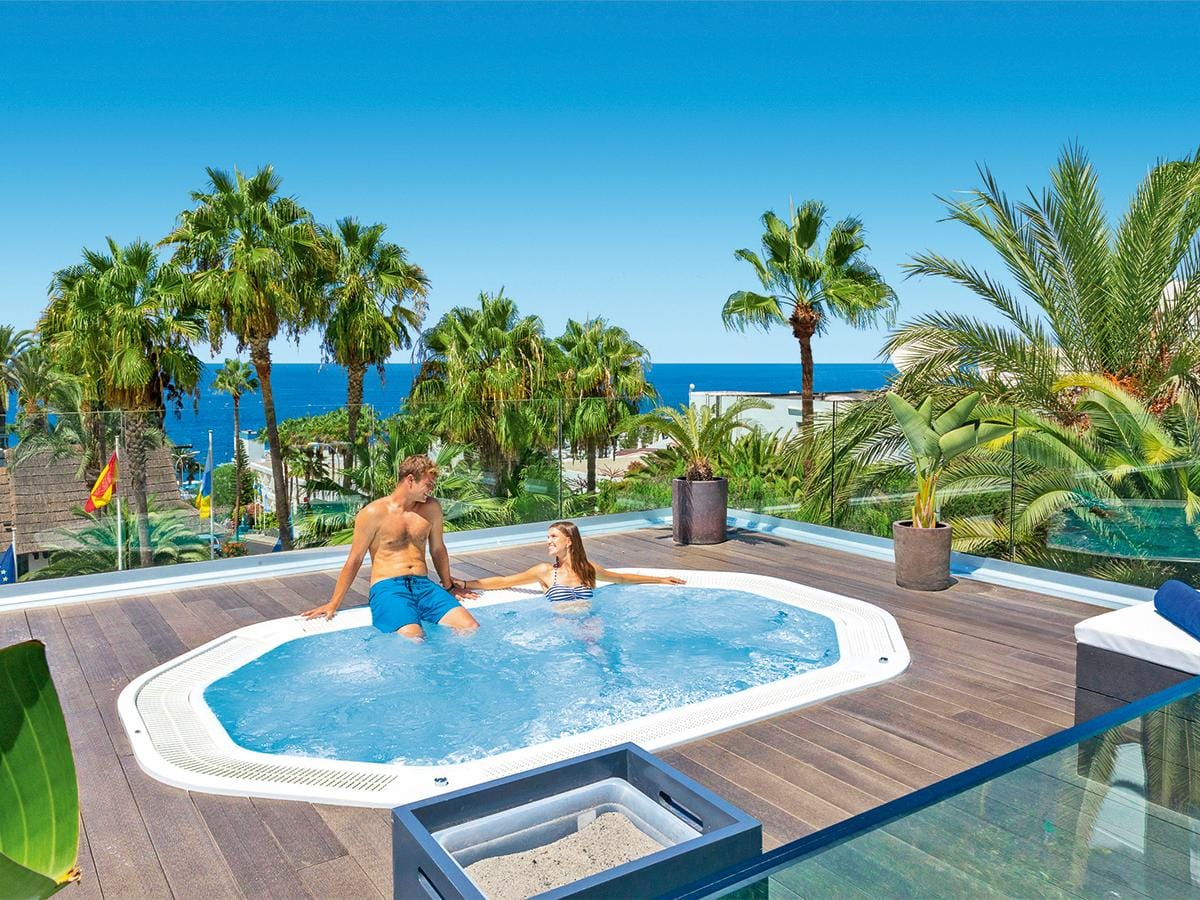 Paar im Jacuzzi auf Hotelterrasse, Meerblick, Palmen, sonniges Wetter, entspannte Atmosphäre.