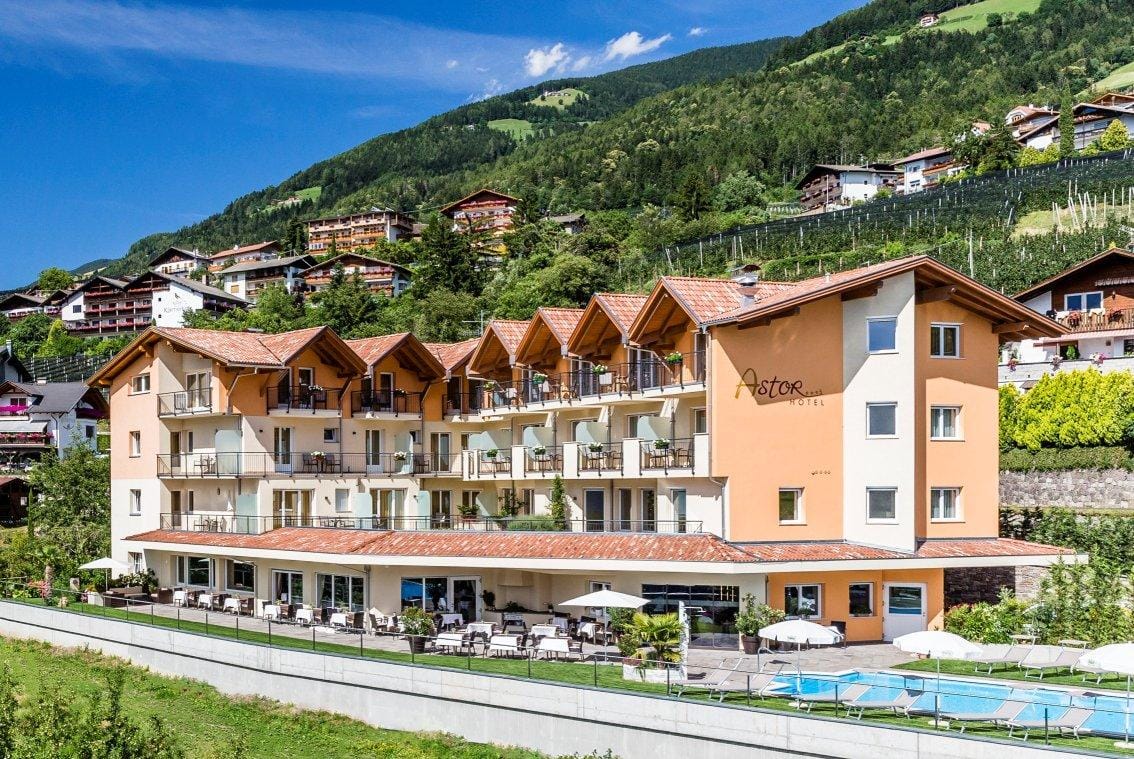 Gelbes Hotel mit Balkonen, Pool im Garten, umgeben von grünen Hügeln und blauen Himmel.