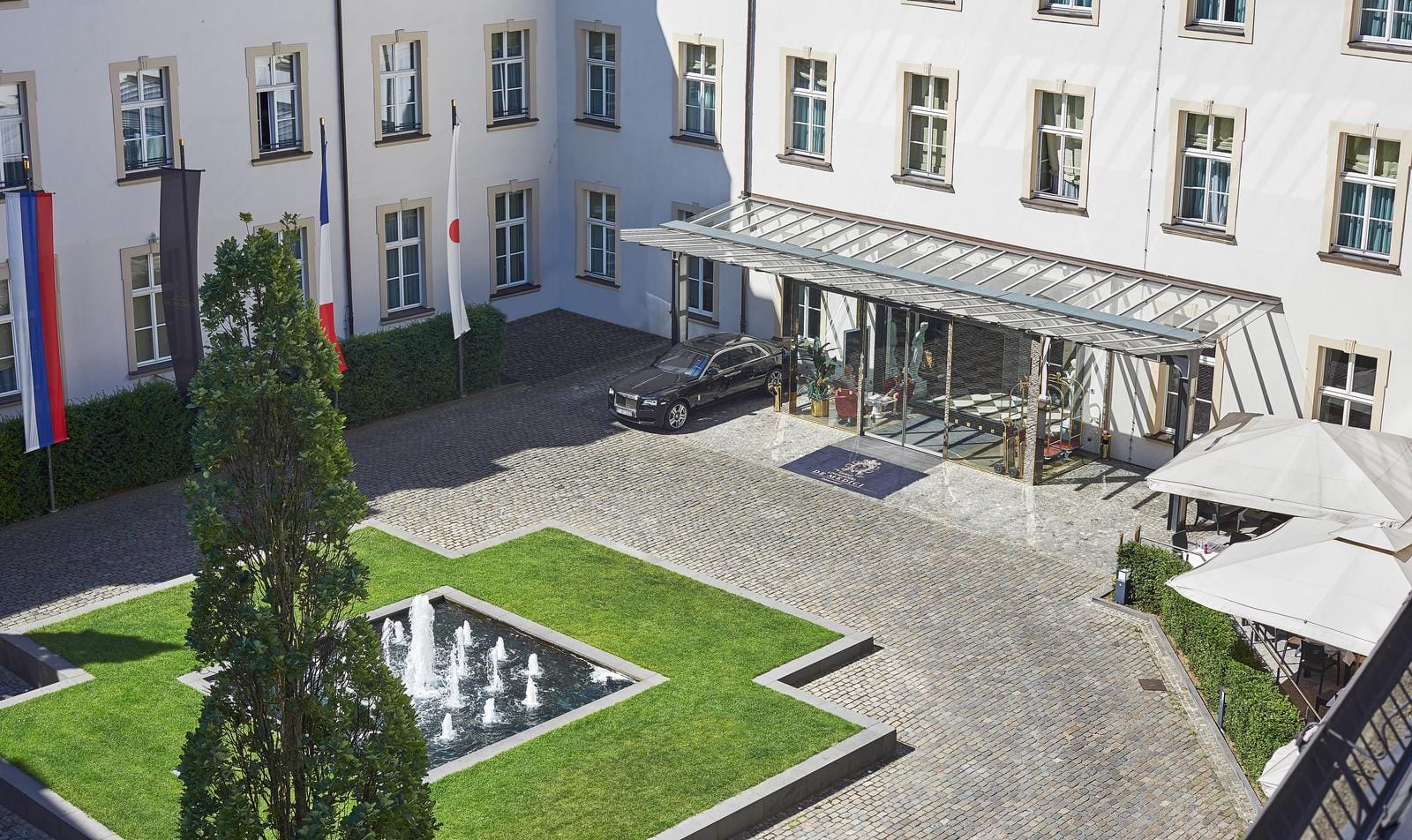 Hoteleingang mit Glaspavillon, gepflegtem Vorplatz und Springbrunnen.