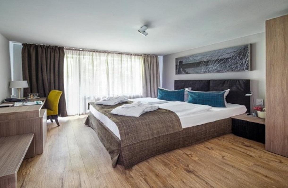 Helles Hotelzimmer mit Doppelbett, Parkettboden, Schreibtisch und bodentiefen Fenstern.