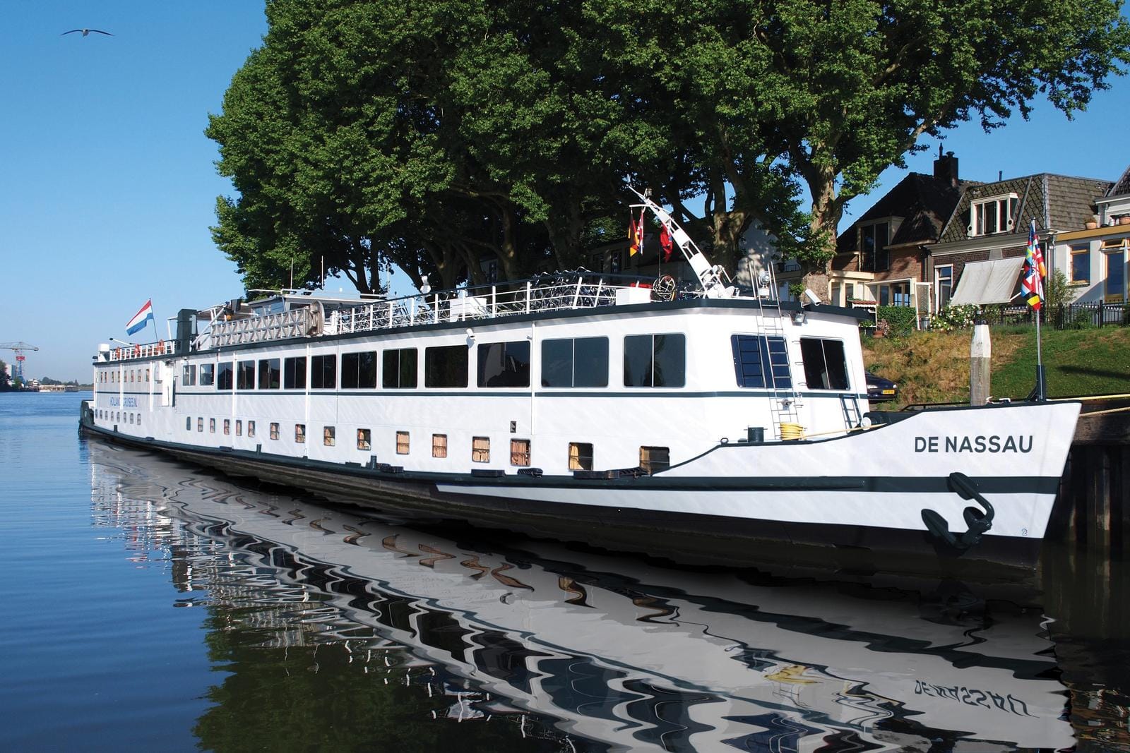Weißes Flussschiff am Ufer, klare Reflexion im Wasser, blaue Flagge, Sommertag.