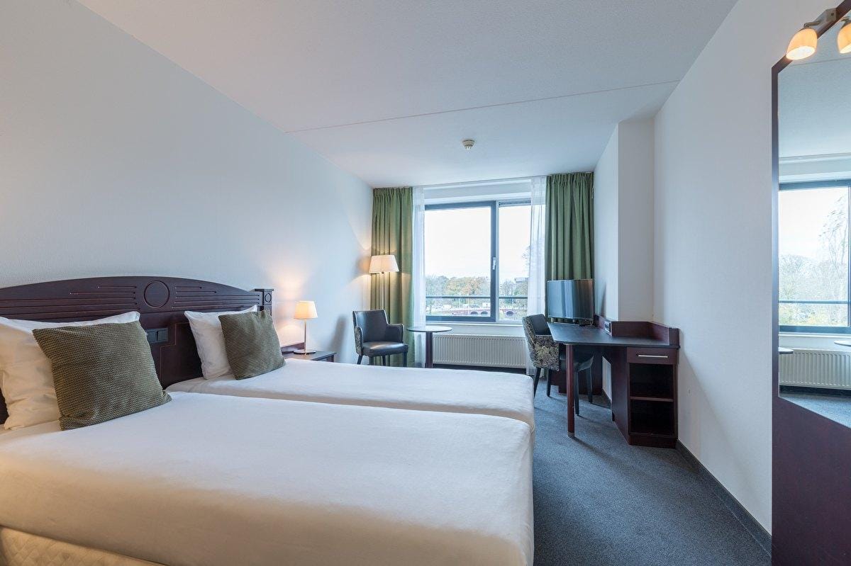 Helles Hotelzimmer mit zwei Einzelbetten, grüne Vorhänge und Schreibtisch mit TV.