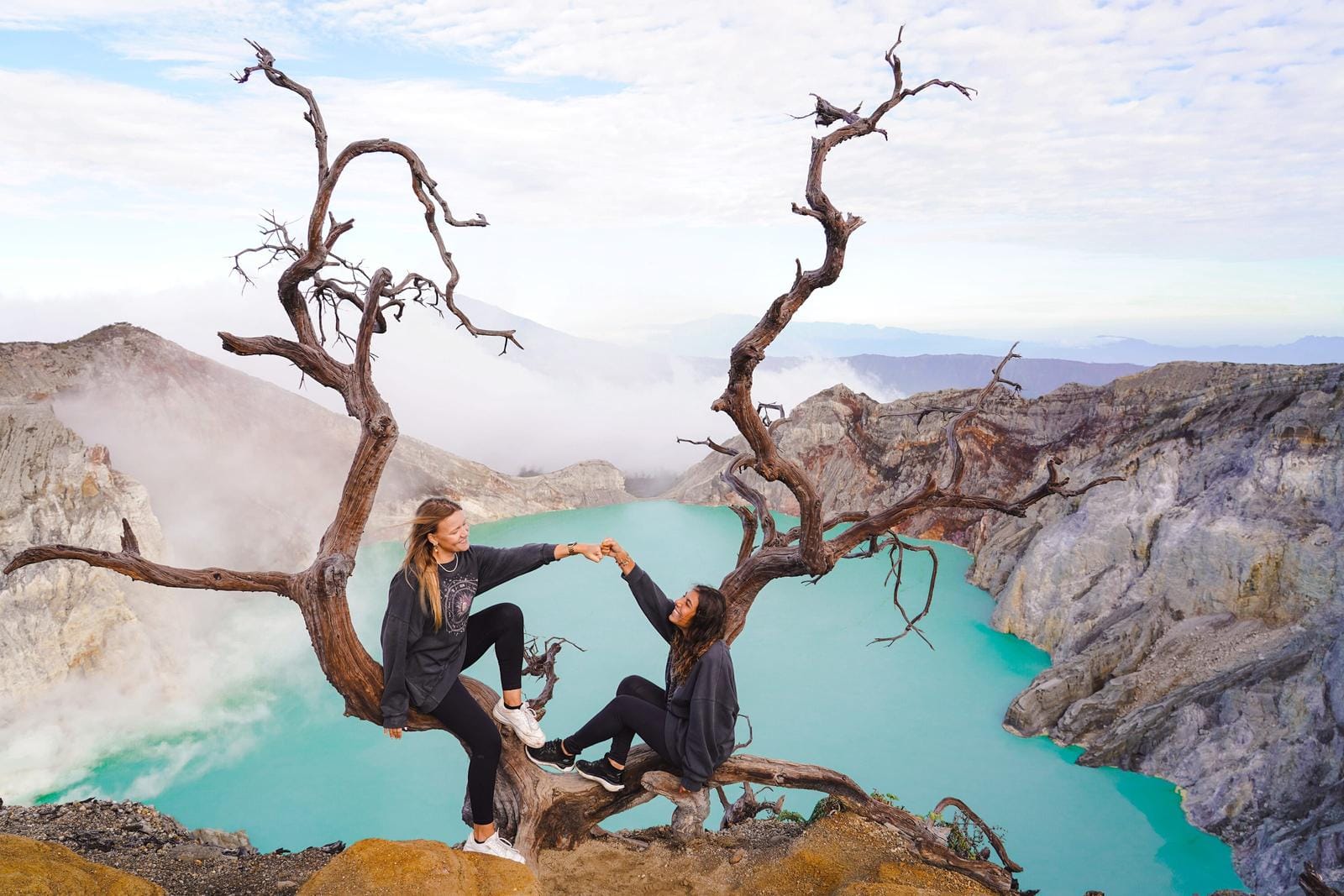 Zwei Personen auf Baumstamm am türkisfarbenen Ijen Kratersee, Indonesien.