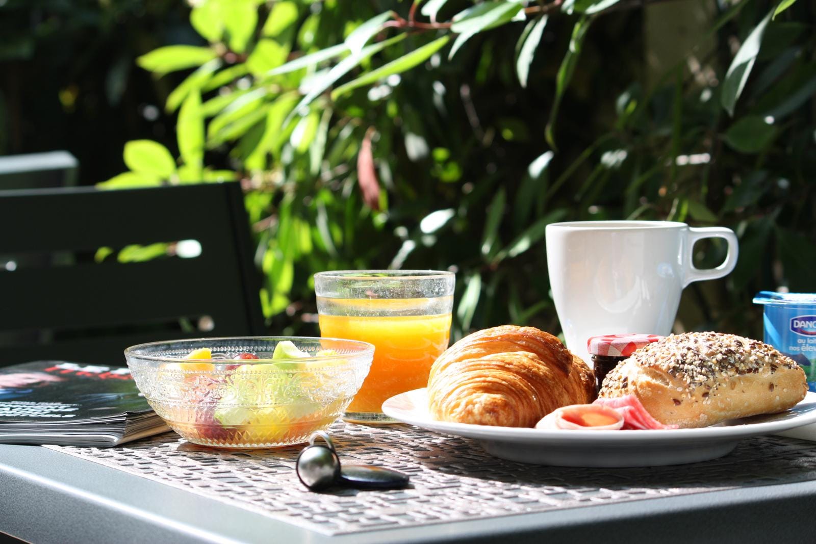 Frühstück auf Terrasse: Croissant, Brötchen, Kaffee, Saft in der Sonne mit grünen Blättern.