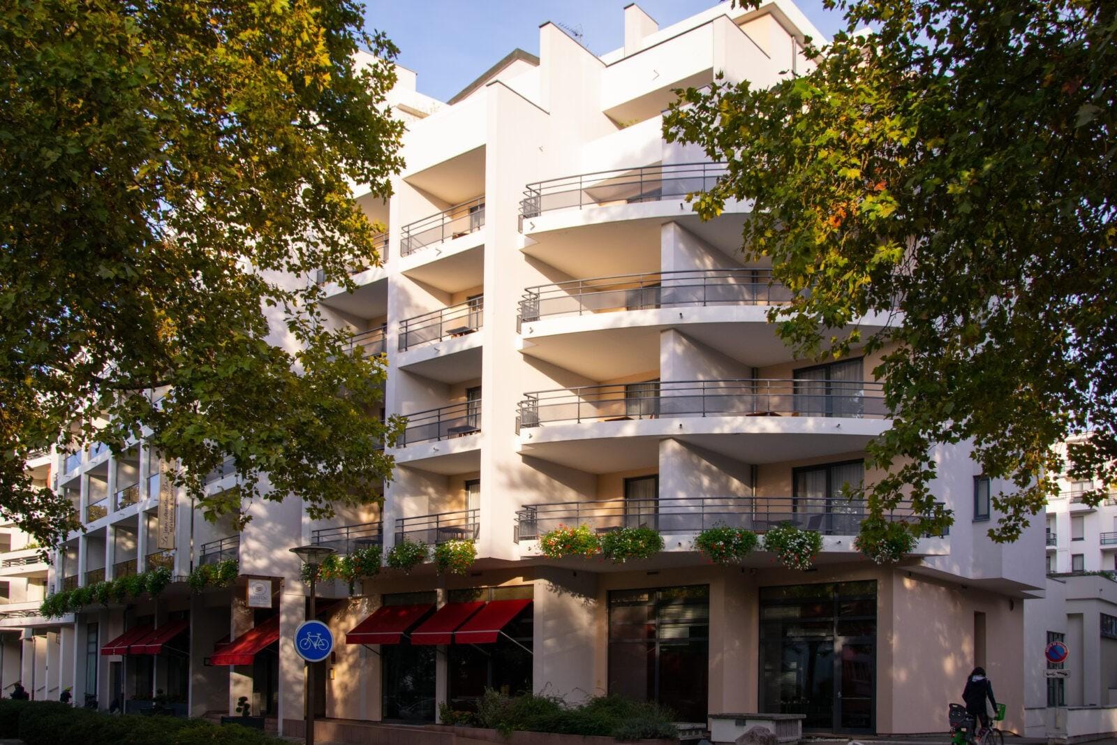 Modernes Hotel in weißem Gebäude mit Balkonen, roten Markisen und Bäumen im Vordergrund.