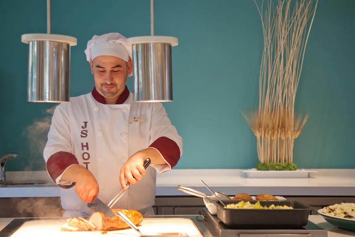 Koch in weißer Uniform schneidet Fleisch unter modernen Lampen vor türkisfarbener Wand.