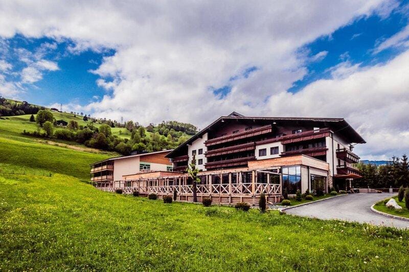 Hotel in idyllischer Berglandschaft, große Terrasse, blau bewölkter Himmel, grüne Wiesen.