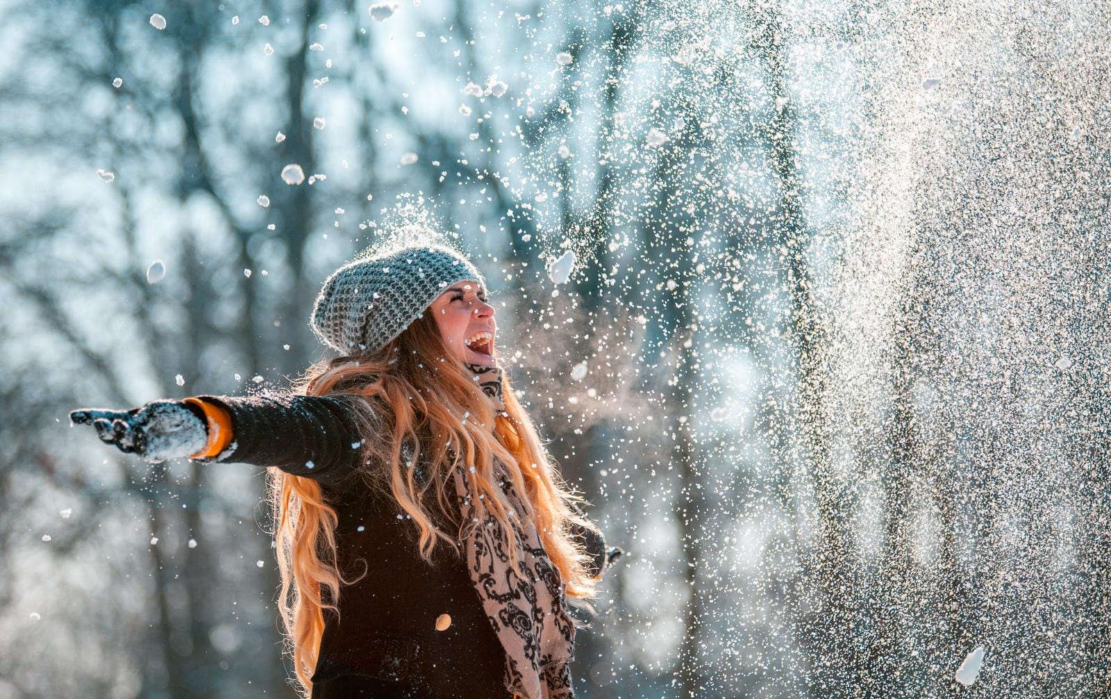 Frau im Wintermantel lacht im Schneefall vor bewaldetem, winterlichem Hintergrund.