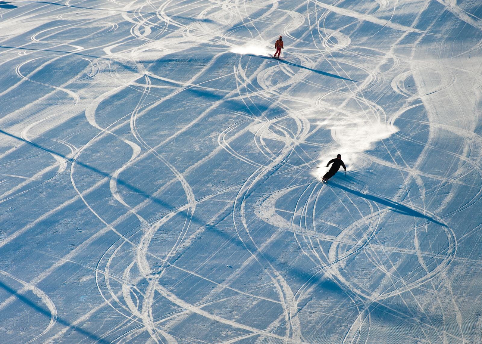 Zwei Skifahrer auf verschneiter Piste mit vielen geschwungenen Spuren im Sonnenlicht.
