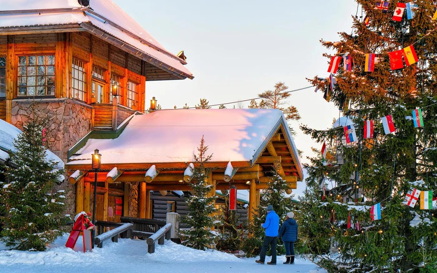 Verschneites Holzhaus in Lappland mit Lichterketten und geschmückten Tannenbäumen.