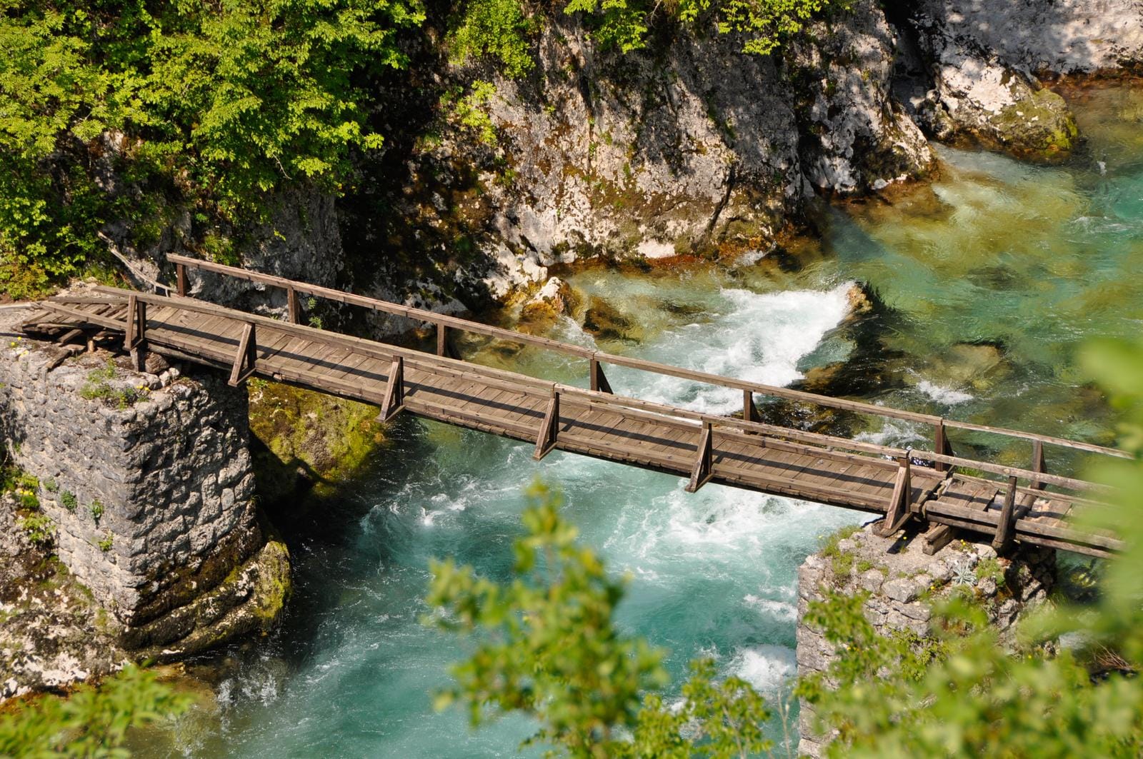 Holzbrücke über türkisfarbenem Gebirgsfluss, umgeben von grüner Vegetation.