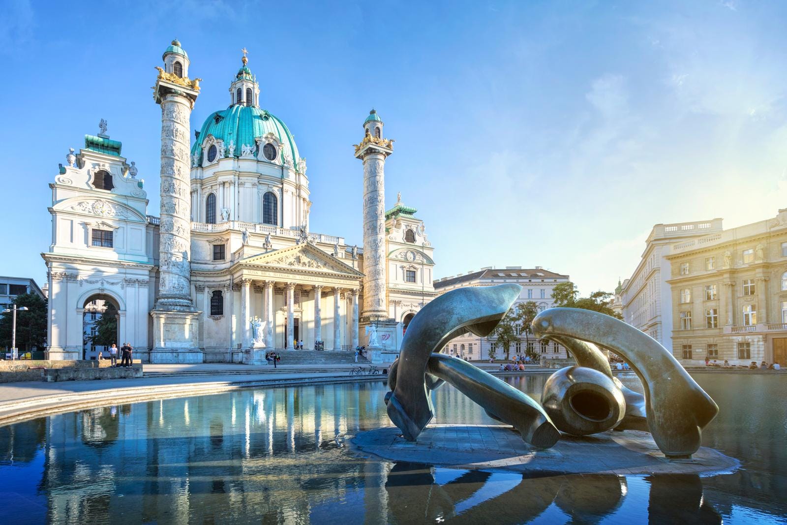 Karlskirche in Wien mit blauem Himmel, reflektiert im Wasser, moderne Skulptur vorne.