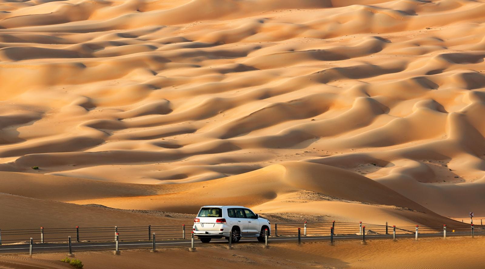 Wüstenstraße mit weißem Auto, umgeben von sanften Sanddünen, in goldener Abendsonne.