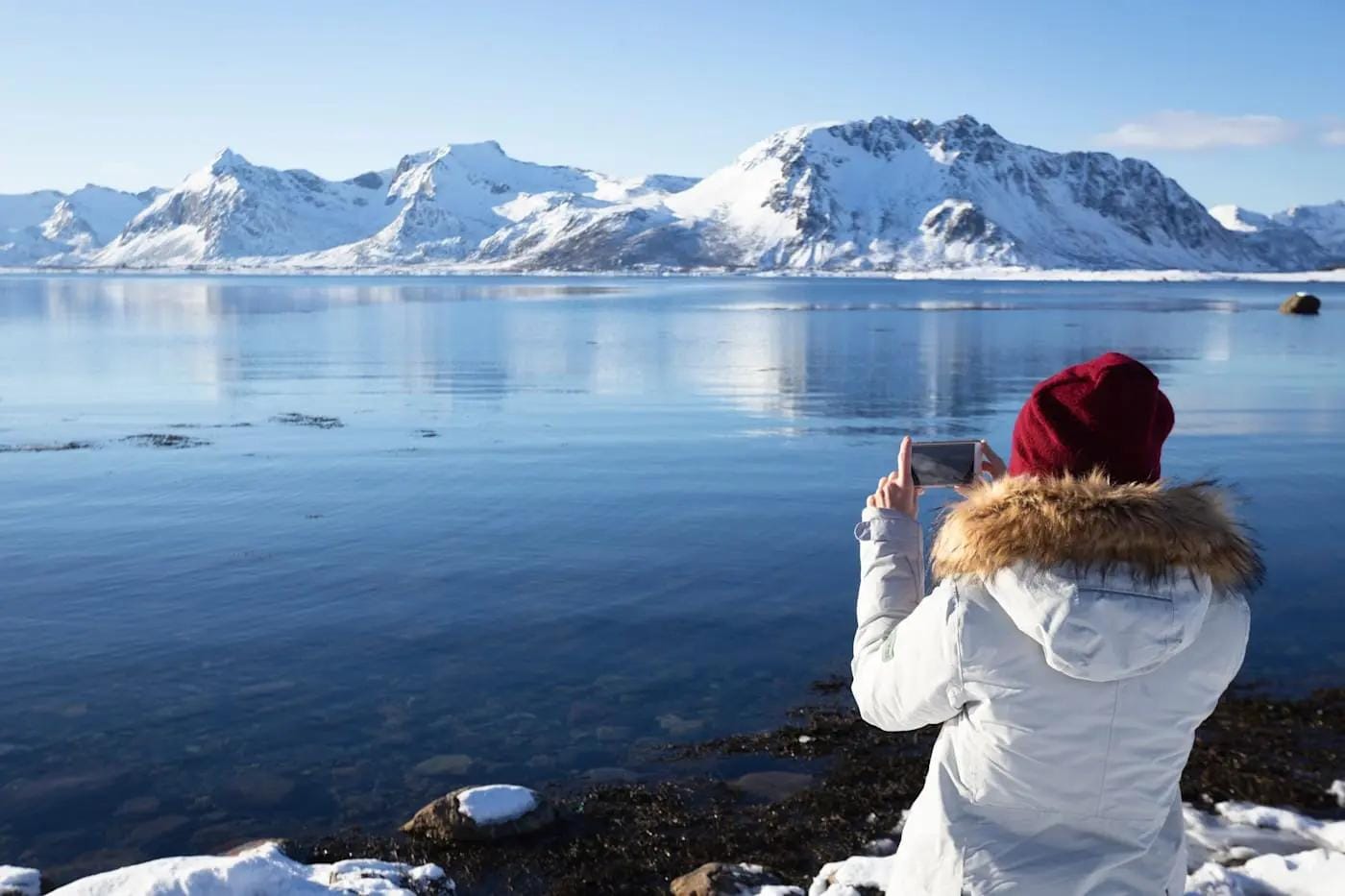 Person in Winterkleidung fotografiert verschneite Berge am Wasser in Norwegen.