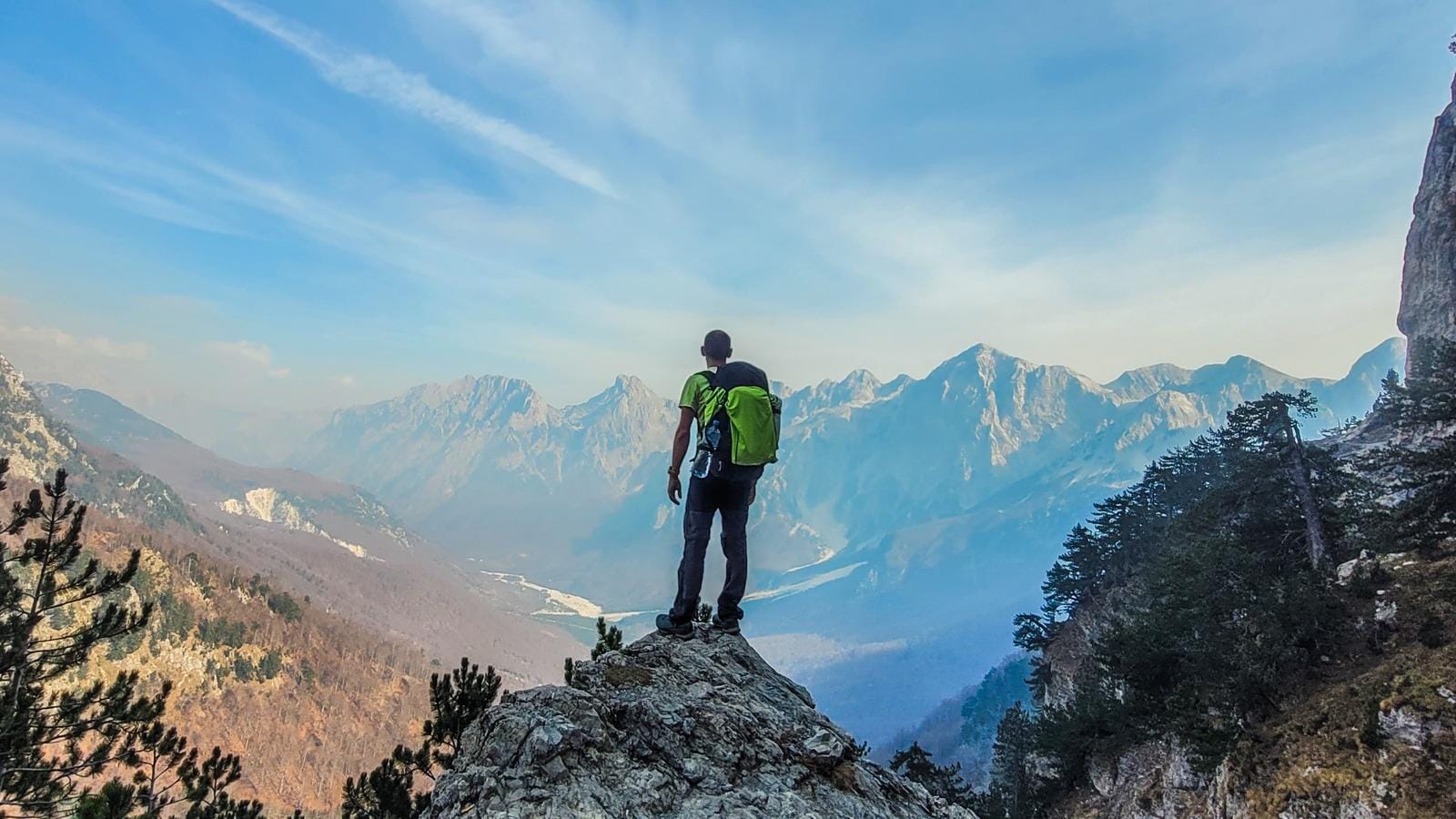Mann mit grünem Rucksack blickt auf majestätische, bewaldete Gebirgskette unter blauem Himmel.