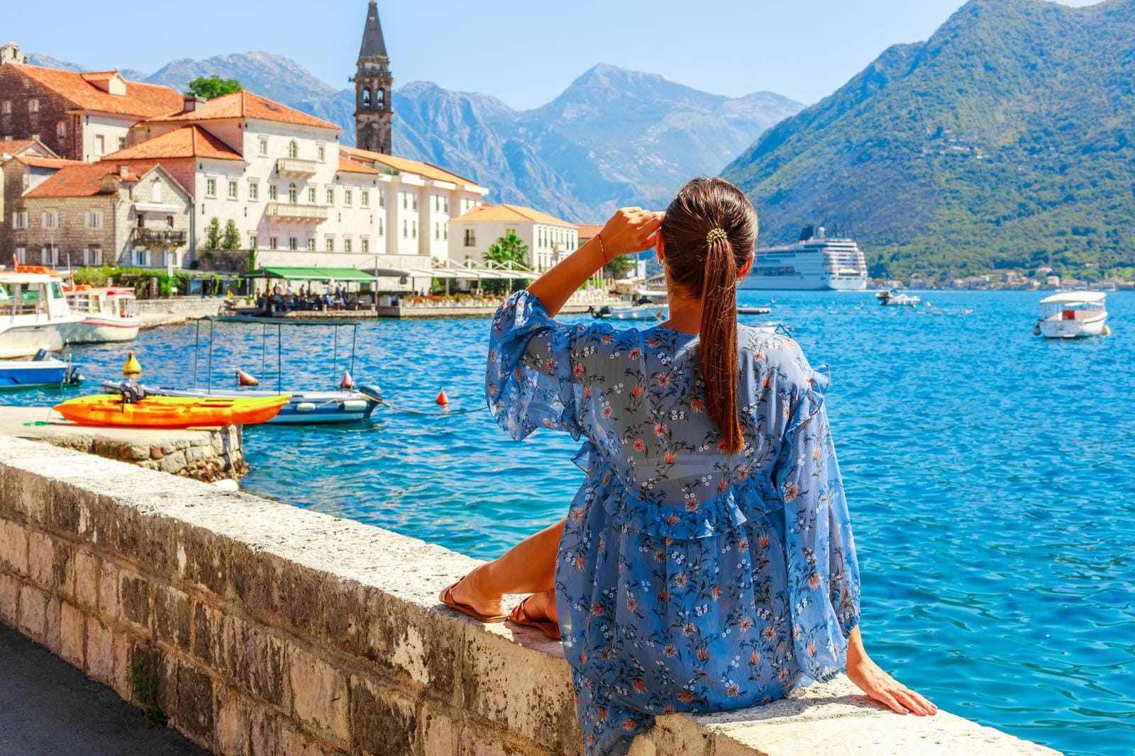 Frau sitzt am Wasser in Perast, Montenegro, mit Bergkulisse und Booten.