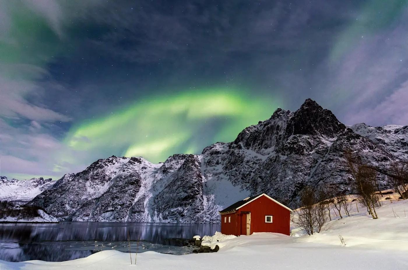 Nordlichter über schneebedeckten Bergen und rotem Haus in Norwegen.