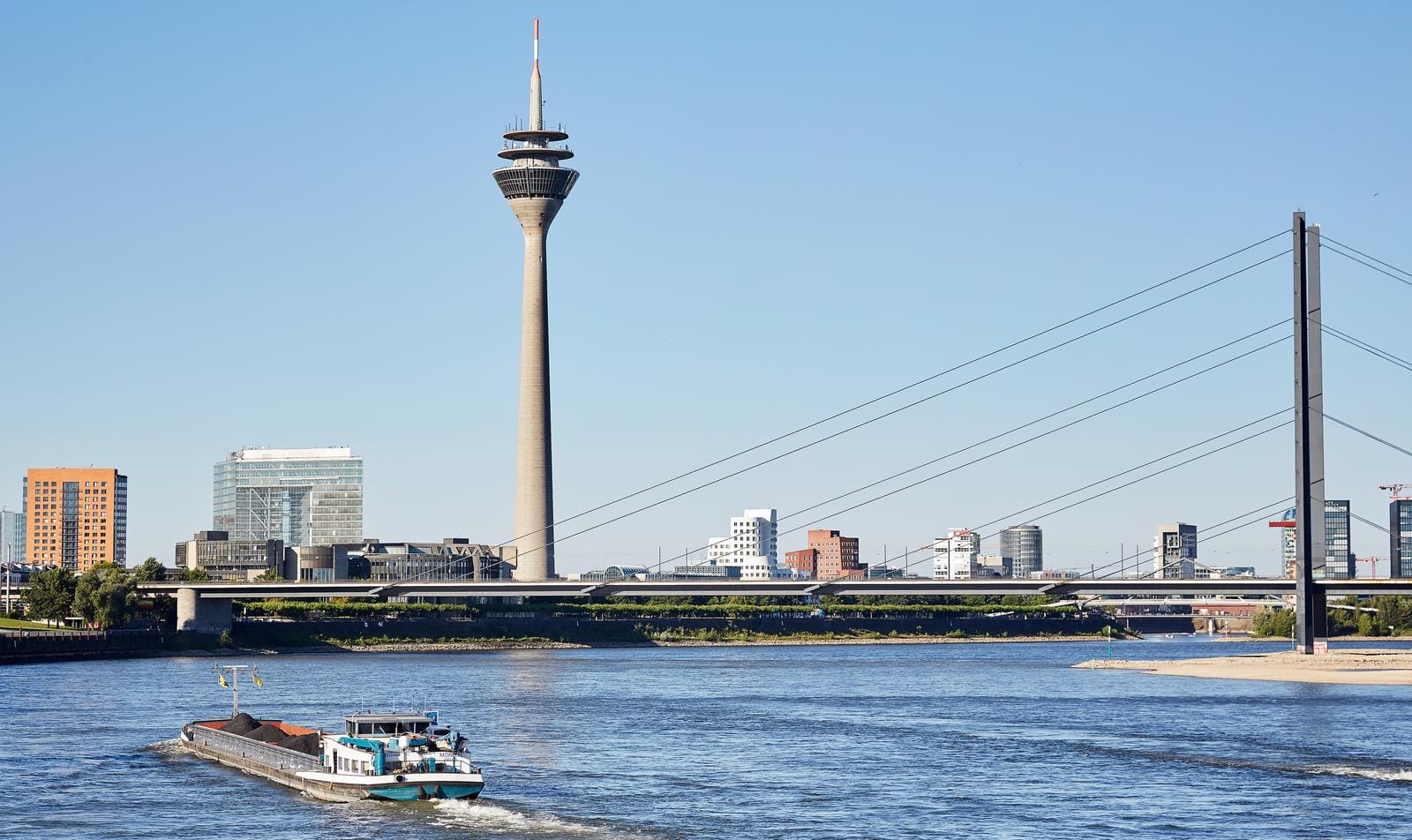 Rheinturm Düsseldorf mit Rheinufer, blauem Himmel und vorbeifahrendem Frachtschiff.