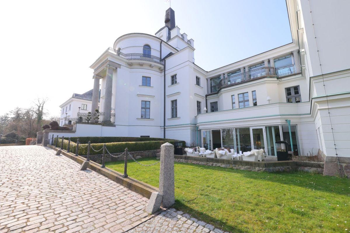 Weiße Fassade eines eleganten Hotels mit Terrasse und Kiesweg bei sonnigem Wetter.