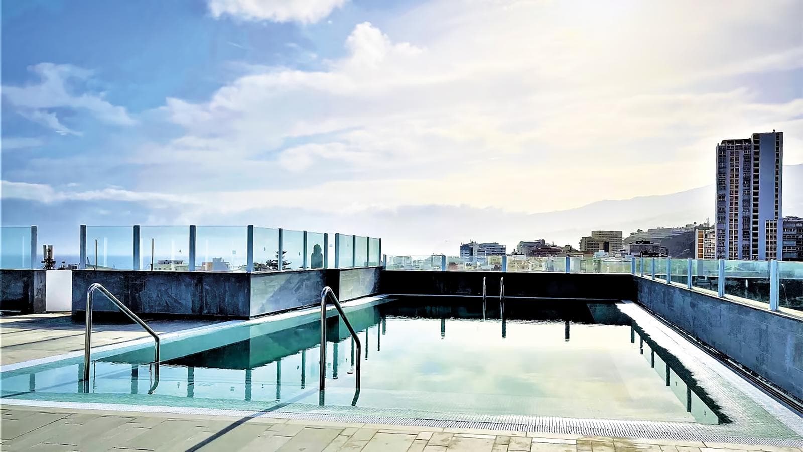 Dachpool mit Stadtblick, gläserne Geländer, Himmel teils bewölkt, moderne Gebäude.