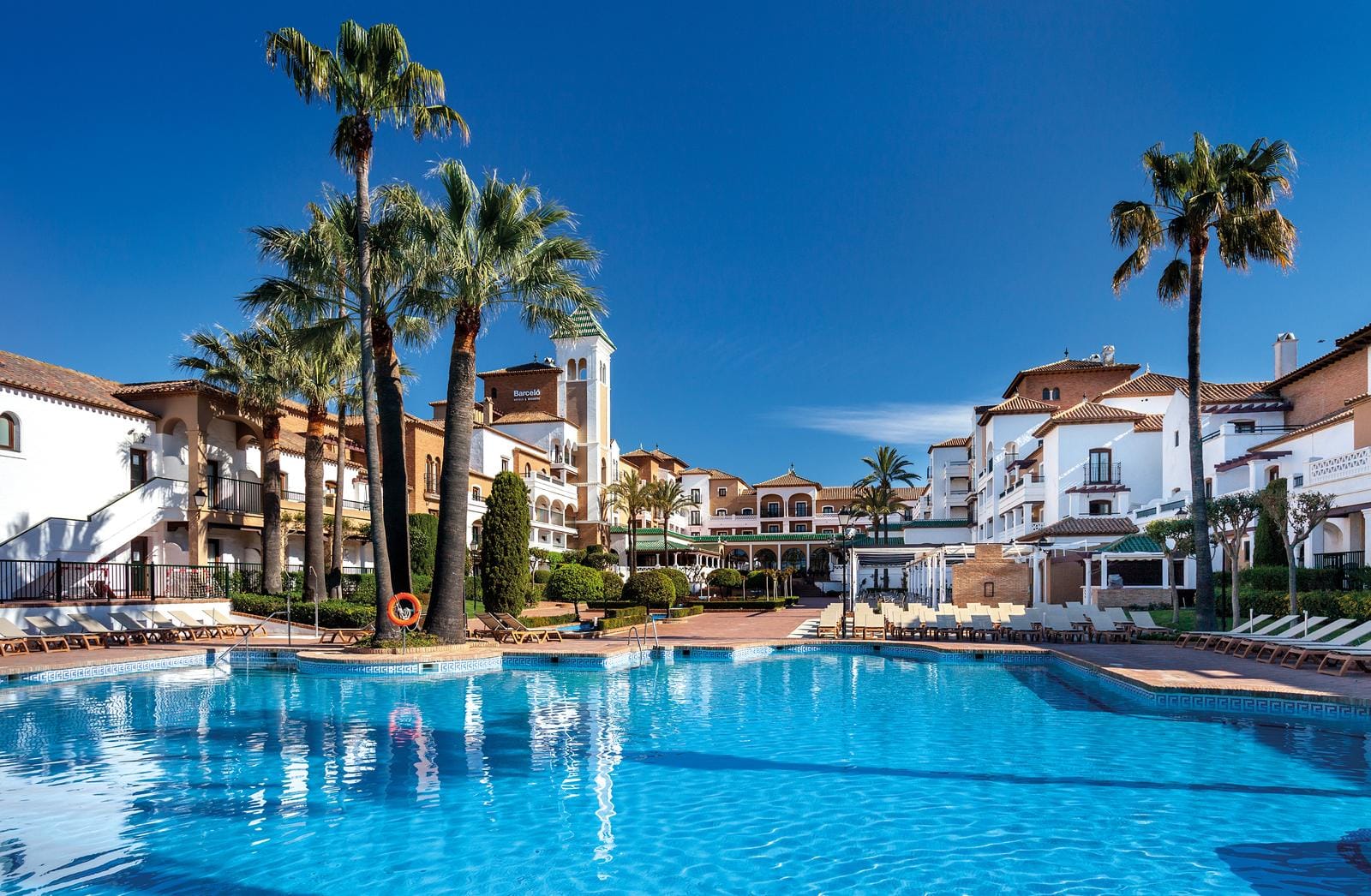 Sonniger Poolbereich in einem spanischen Küstenresort mit Palmen und mediterraner Architektur.