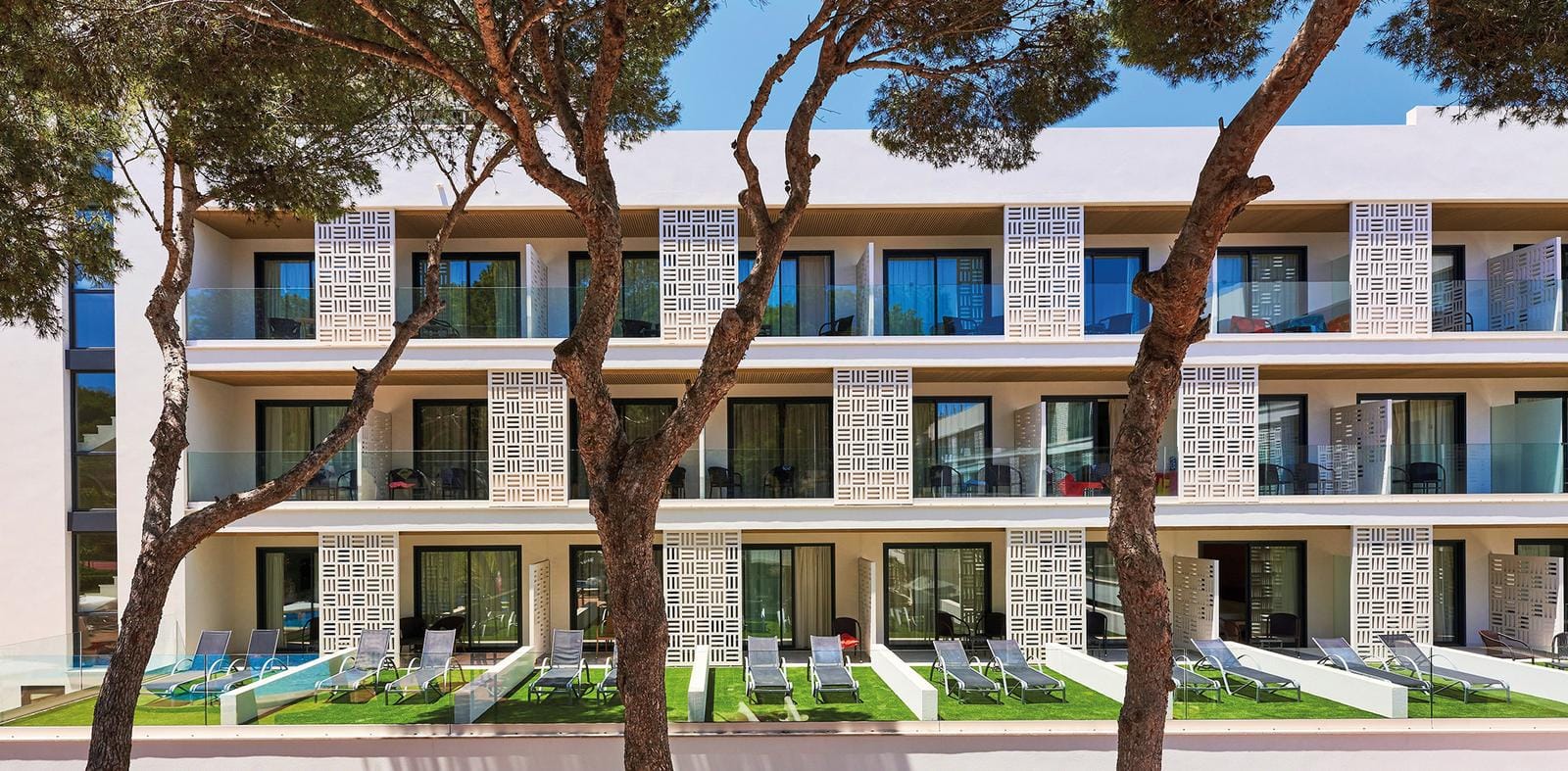 Modernes Hotelgebäude mit Balkonen, Glasgeländer und Sonnenliegen auf grünem Rasen.